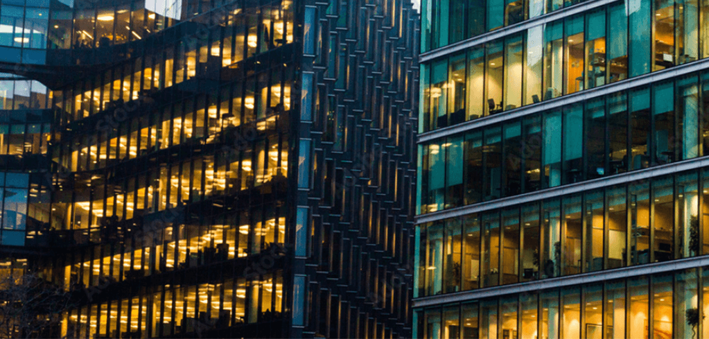 Two illuminated glass office buildings at dusk, glowing with warm golden and teal lights across multiple floors.