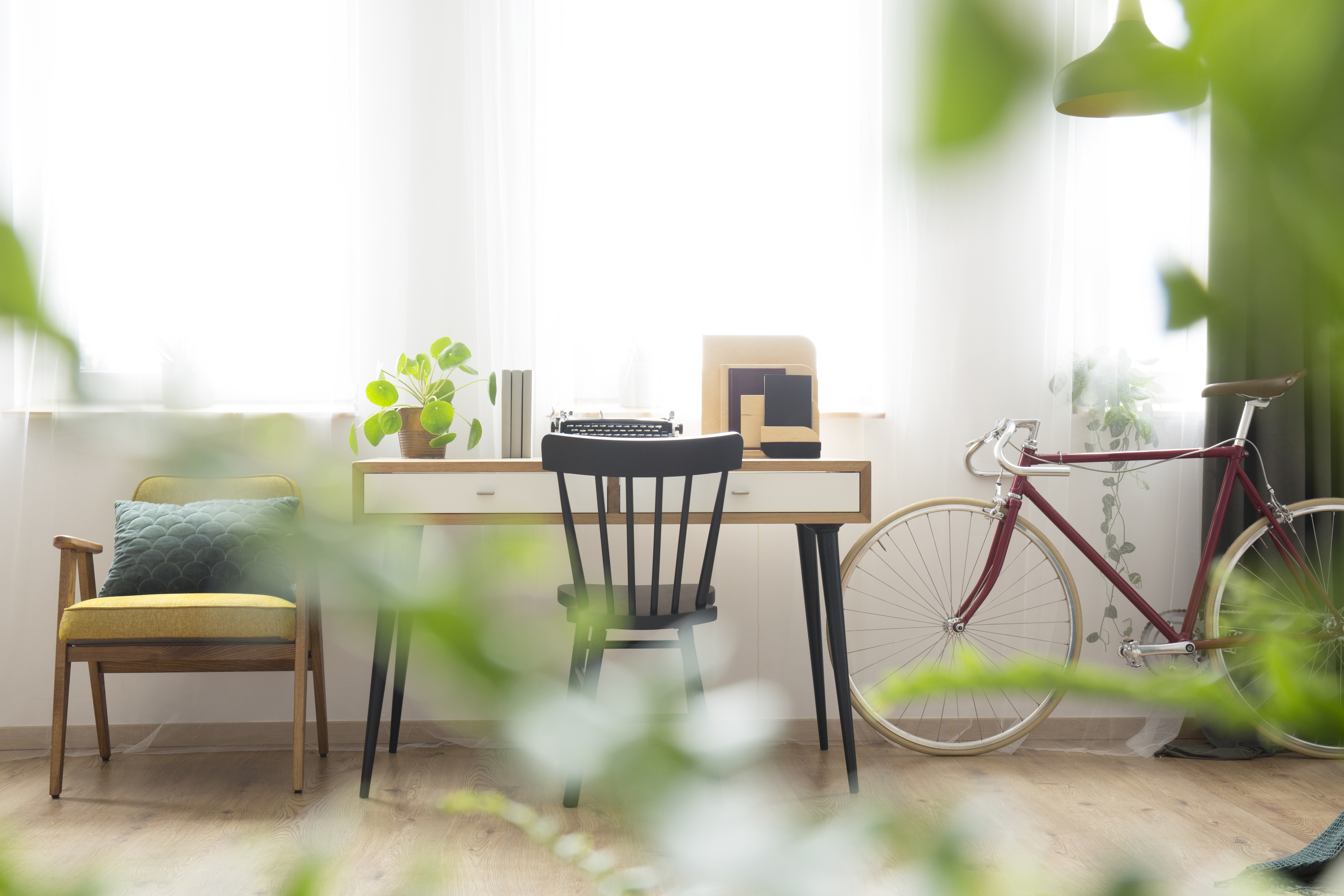 Bright home office with wooden desk, typewriter, black chair, yellow cushioned seat, and red bicycle against white walls.