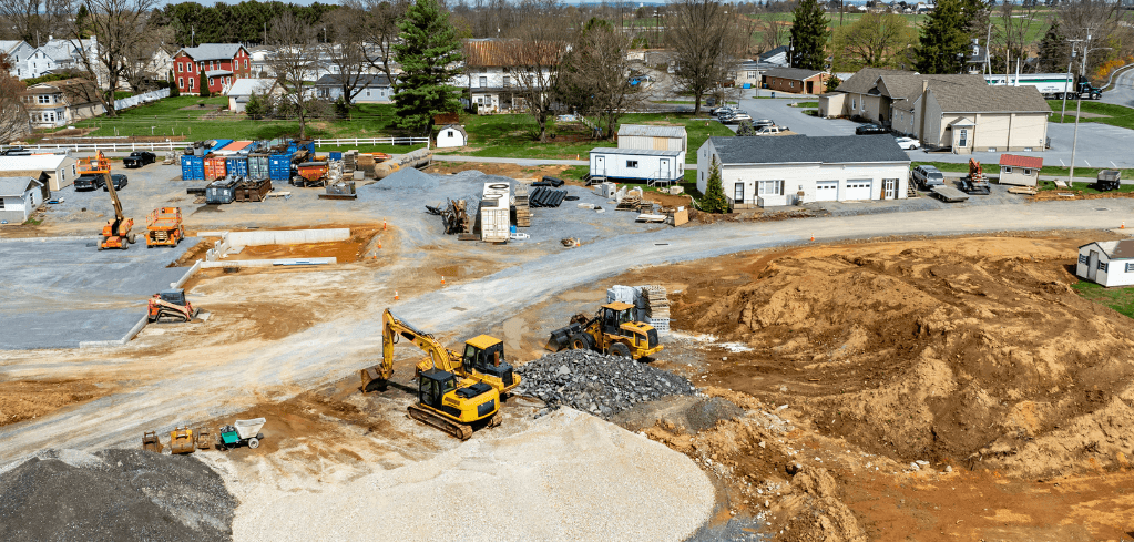 Aerial view of an active construction site with yellow excavators, dirt mounds, gravel, and buildings in a suburban area.