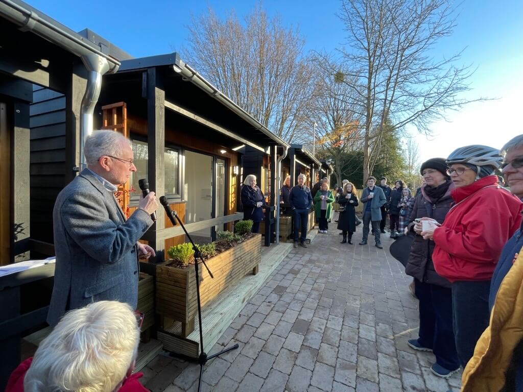 An elderly man in a gray suit speaks into a microphone outdoors at a gathering, with a crowd listening near a modern wooden building.