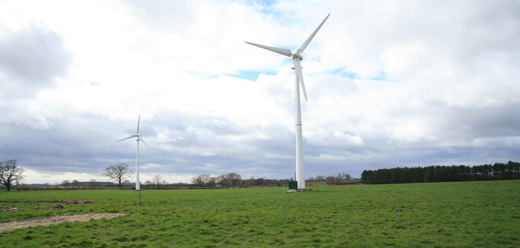 Two white wind turbines standing in a green field under a cloudy sky with patches of blue visible.