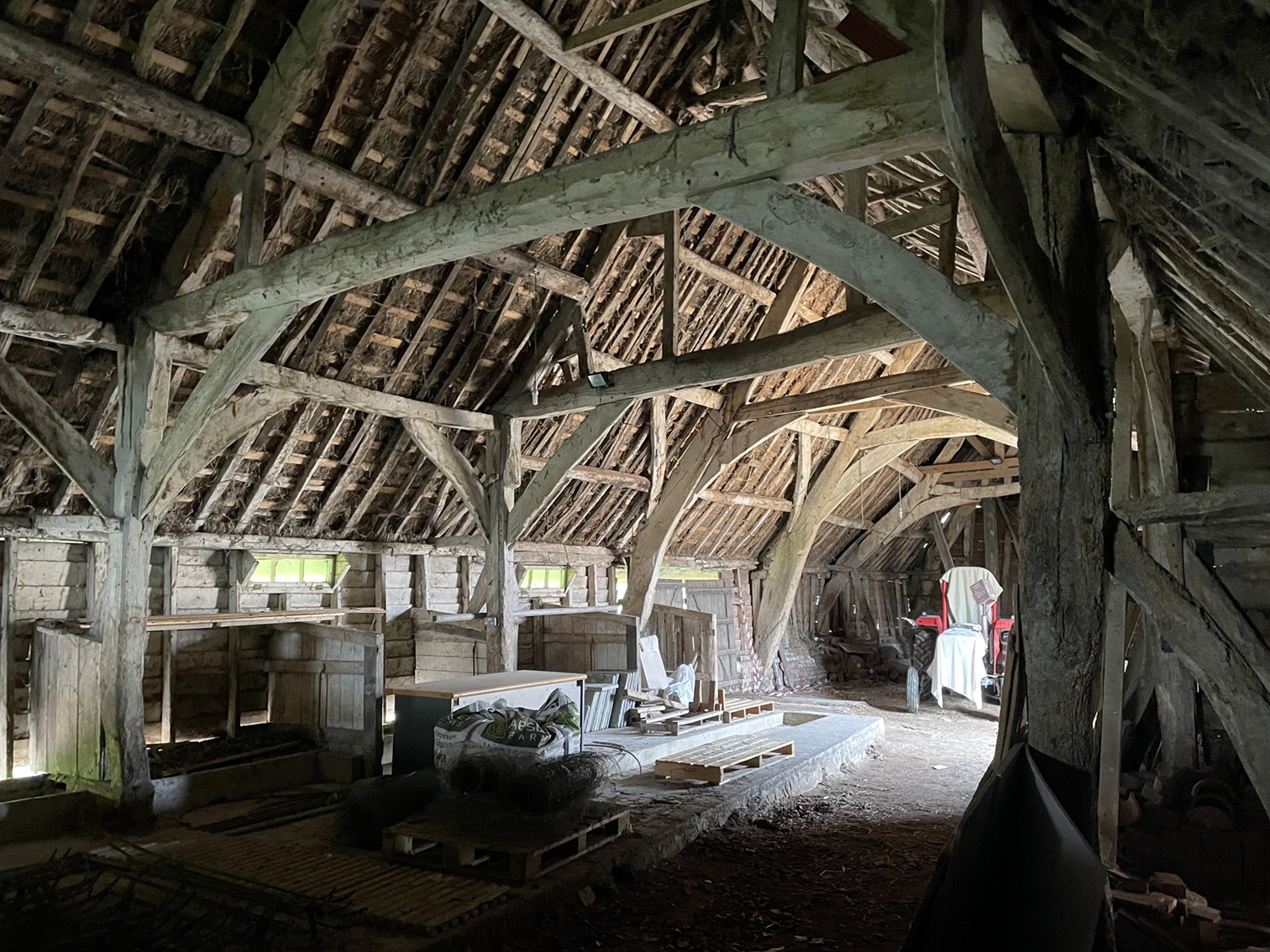 Interior of an old wooden barn with exposed timber beams, scattered storage items, and natural light filtering through windows.