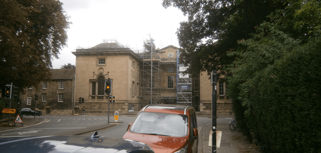 A historic stone building under renovation with scaffolding, traffic lights, and cars at a street intersection surrounded by trees.