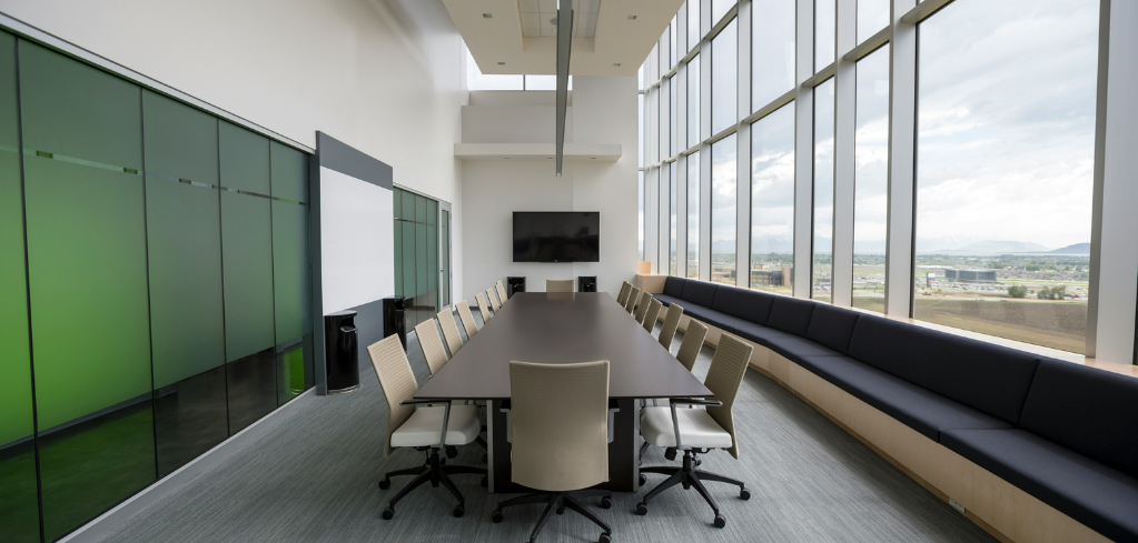 A modern conference room with a long dark table, beige chairs, a wall-mounted TV, green glass panels, and floor-to-ceiling windows.