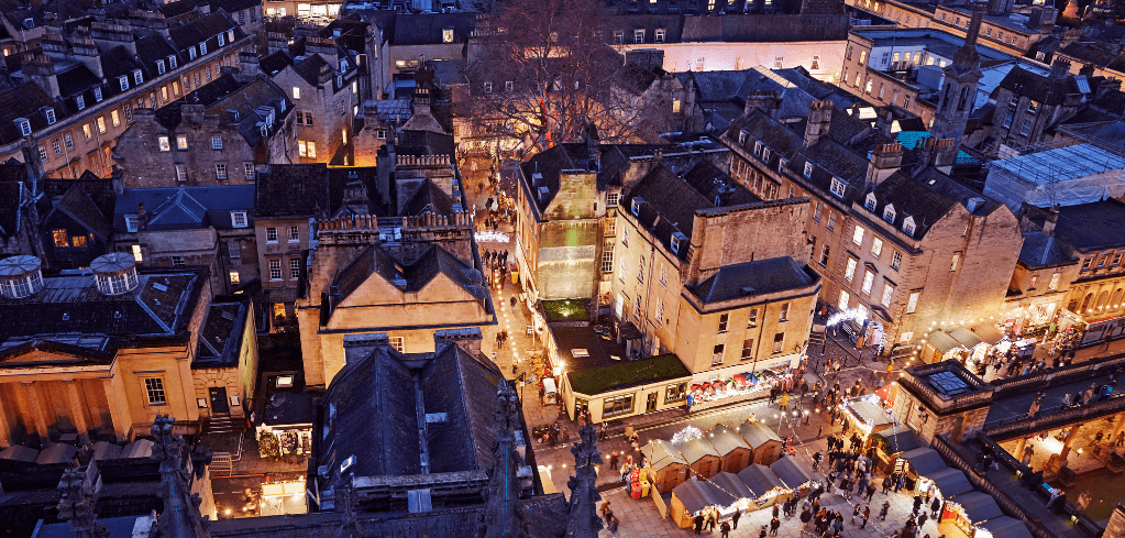 Aerial night view of Bath city centre with illuminated stone buildings and a bustling Christmas market below.