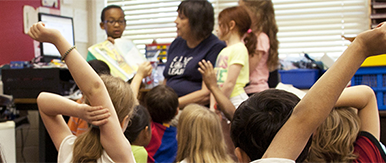 A classroom scene with young children sitting on the floor, raising their hands while a teacher leads an activity at the front of the room.