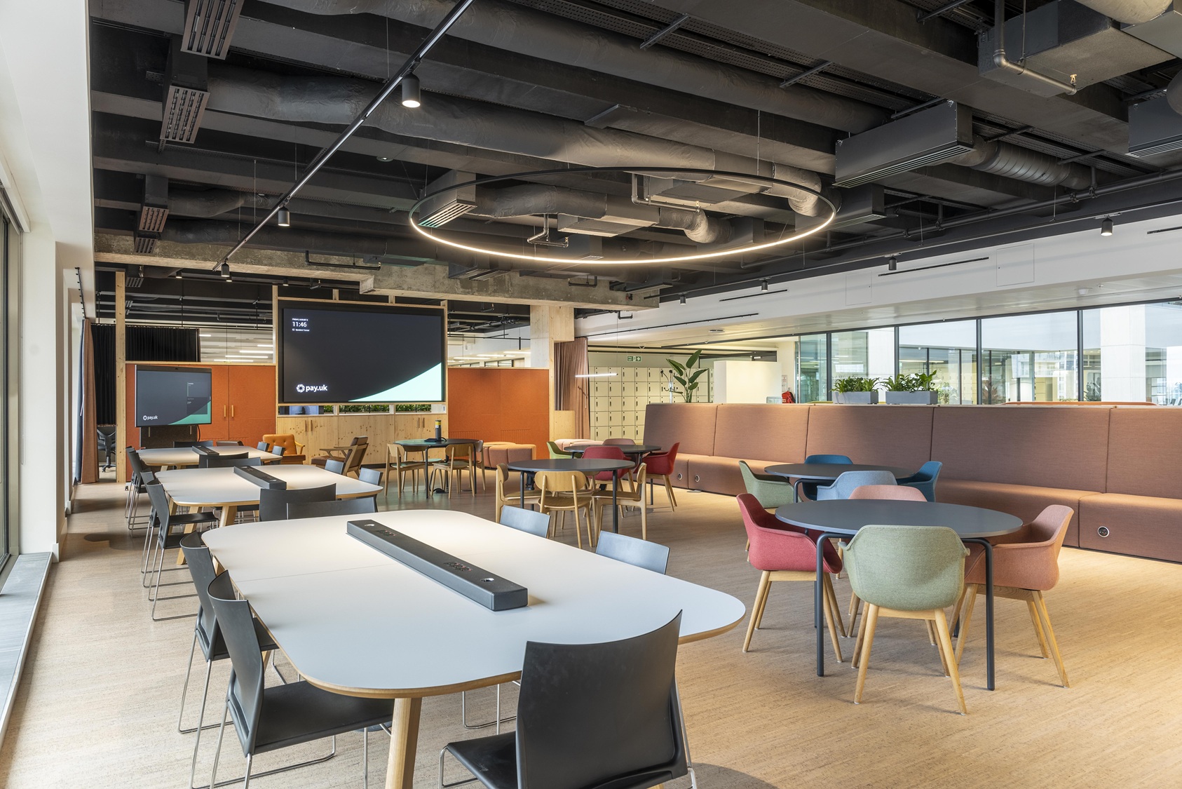Modern office space with colorful chairs, conference tables, orange accent walls, and a large display screen in an open layout.