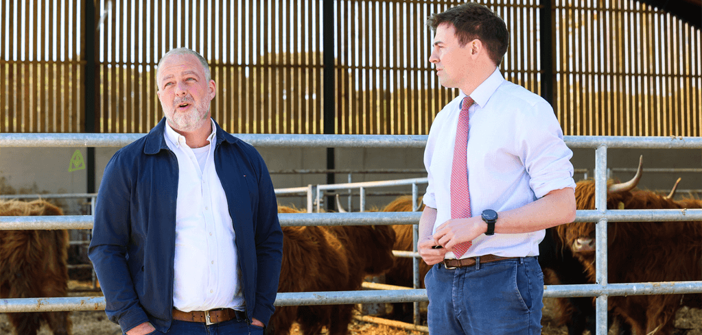 Two men in conversation at a cattle farm, with Highland cows visible in metal pens behind them.