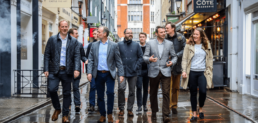 Group of professionals walking together on a wet city street with shops and restaurants visible in the background.