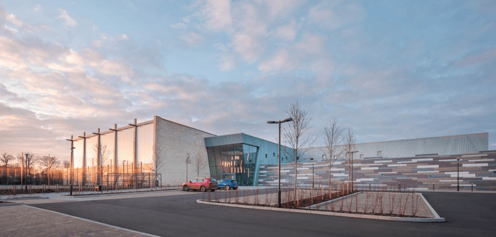 Modern sports facility exterior at dusk with teal glass entrance, two parked cars, bare trees, and a pastel cloudy sky.