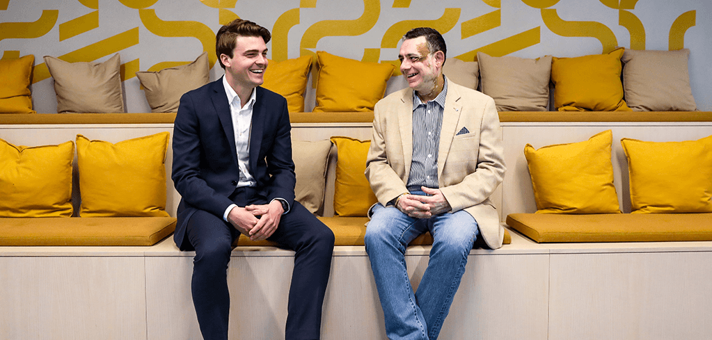 Two men in business attire having a conversation while seated on yellow cushioned benches with matching wall design.
