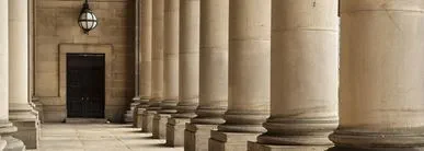 A row of large stone columns forming a classical architectural colonnade, with a dark doorway and a hanging lantern at the end.