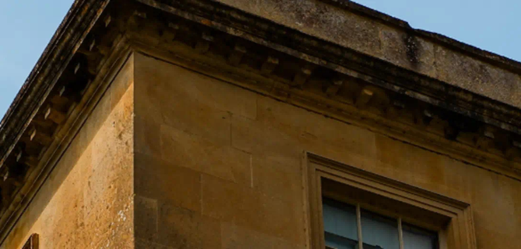 Corner of a historic stone building with beige limestone walls, ornate cornice, and partial window view against blue sky.