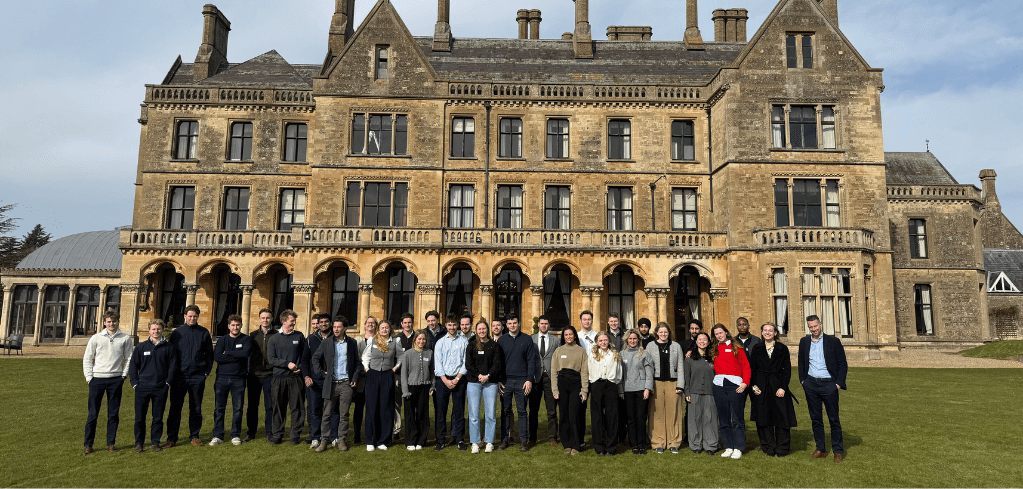 Large group gathered on lawn in front of historic stone mansion with ornate Victorian architecture and multiple chimneys.