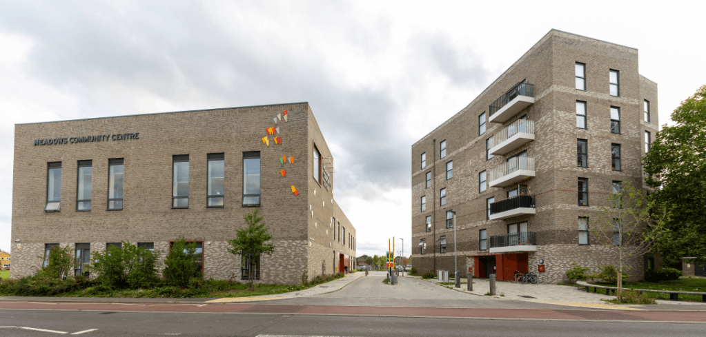 Meadows Community Centre brick building beside a multi-story residential block, separated by a pedestrian pathway under a cloudy sky.