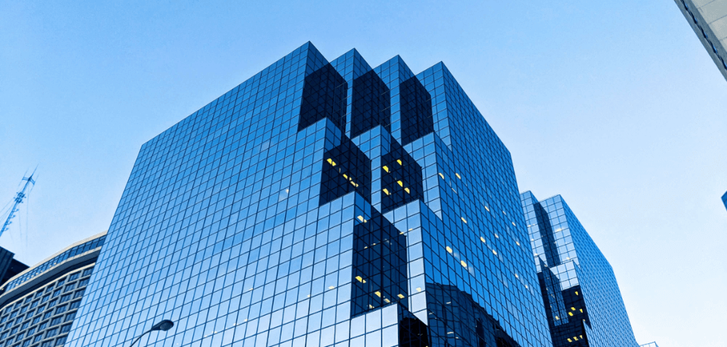 Modern blue glass skyscraper with reflective facade against clear sky, few office lights visible inside.