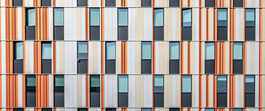A close-up view of a modern building façade featuring vertical panels in orange, beige, and grey tones, arranged in a repeating pattern with evenly spaced windows.