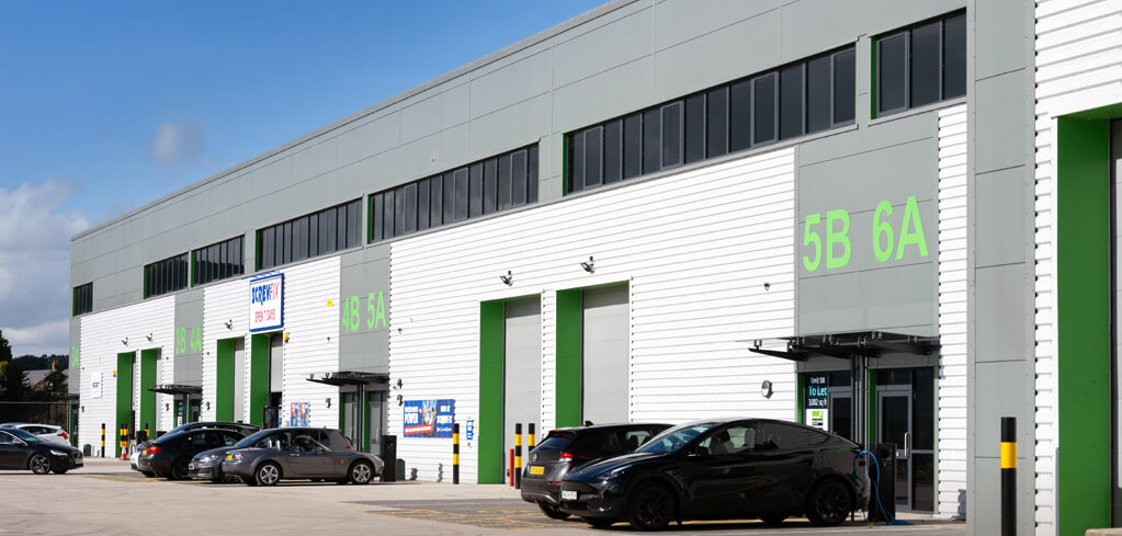Modern industrial units with green-trimmed doors numbered 5B and 6A, cars parked outside on a sunny day.