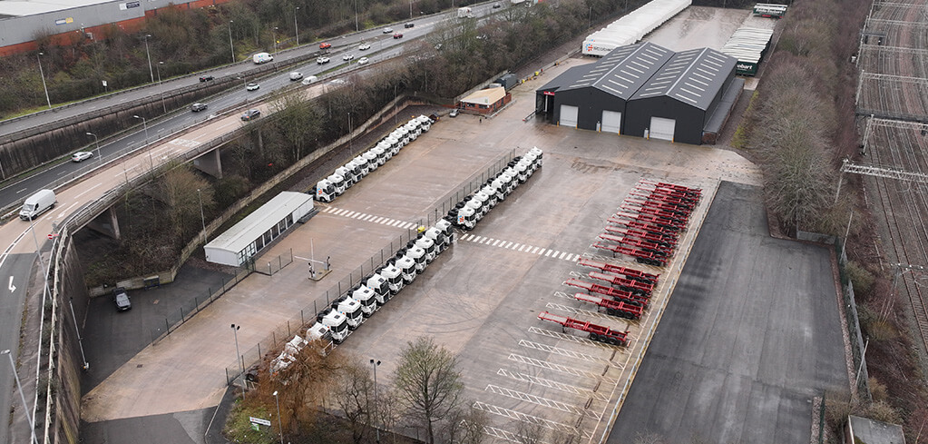 Aerial view of a large truck depot with rows of white cab units and red trailers parked near a dark industrial warehouse beside a motorway.