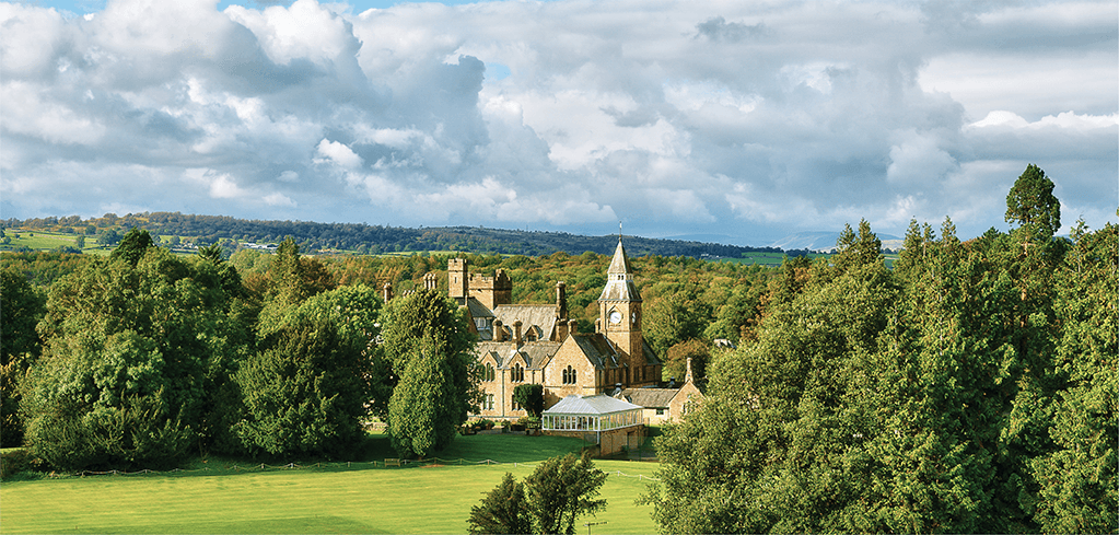 Historic stone building with clock tower nestled among green trees under cloudy sky in rolling countryside landscape.