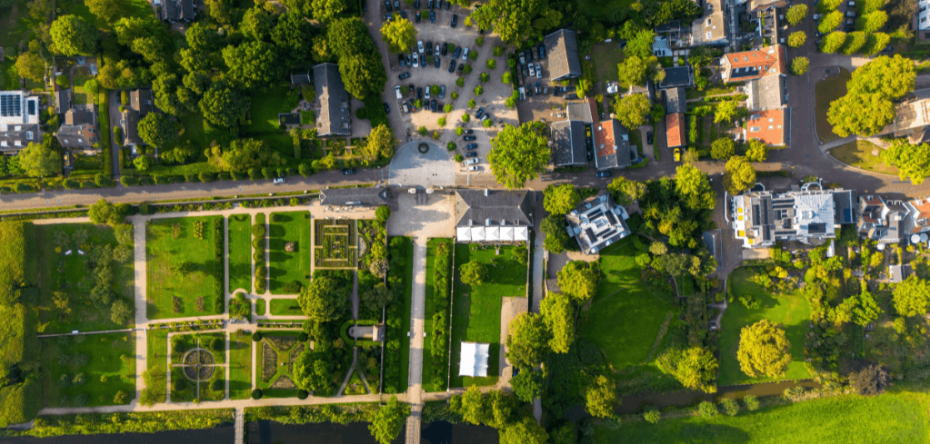Aerial view of a lush green town with formal gardens, tree-lined streets, buildings, and parked cars on a sunny day.