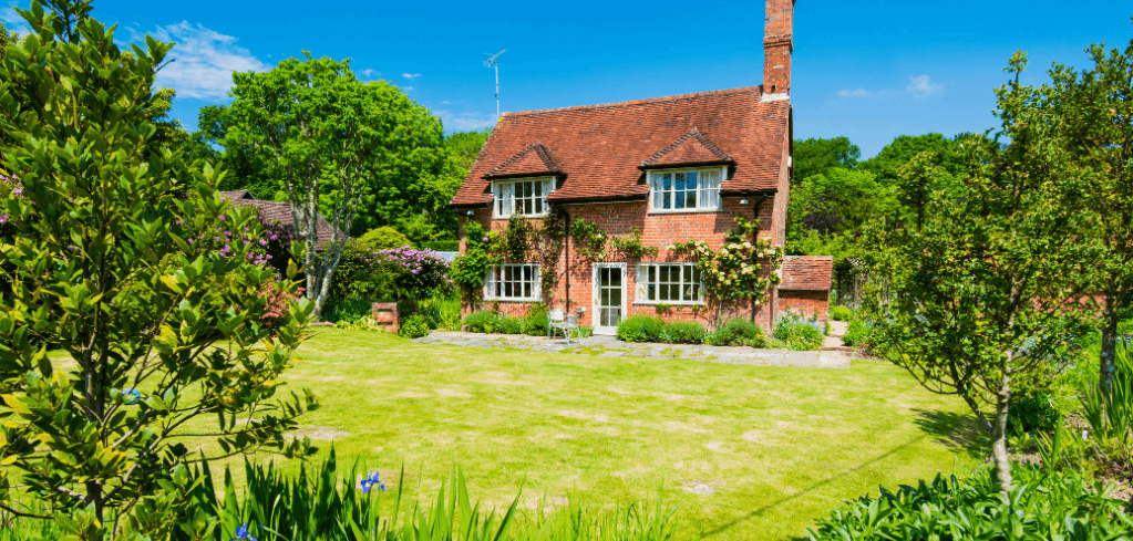 Traditional brick cottage with red tile roof surrounded by lush green garden and lawn on a bright sunny day.