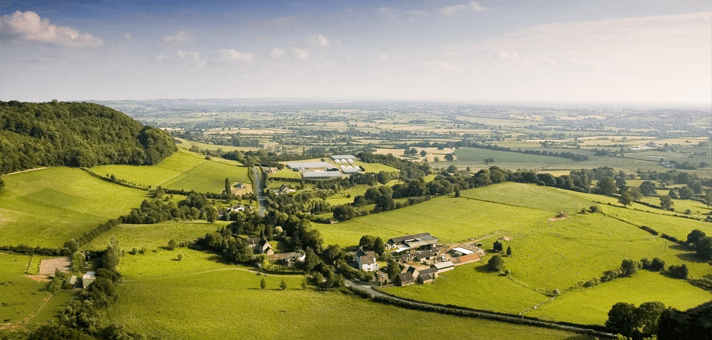 Aerial view of lush green countryside with rolling hills, scattered farm buildings, and patchwork fields stretching to the horizon.