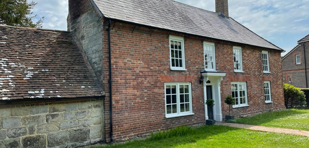 Traditional red brick country house with white-framed windows, slate roof, and stone extension under partly cloudy sky.
