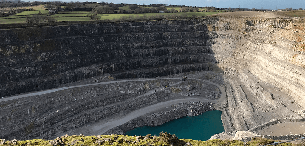 Aerial view of a large open-pit quarry with tiered rock walls, machinery on roads, a teal pool, and green fields above.