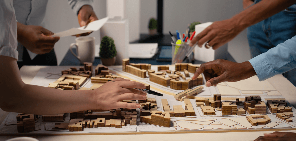 A group of people collaborating around a table with a detailed wooden architectural scale model of a city layout.
