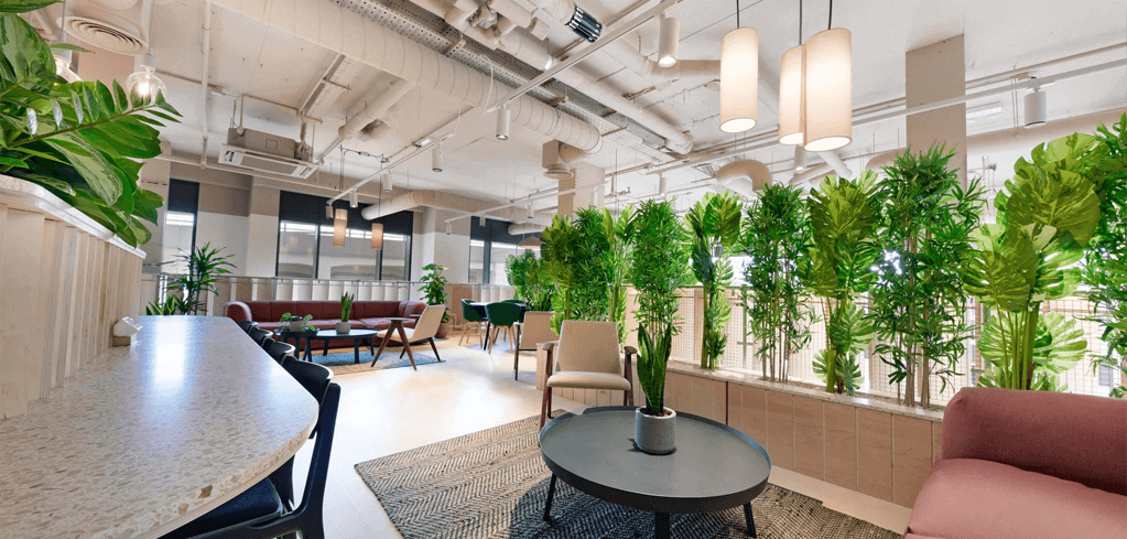 Modern office lounge with abundant indoor plants, pendant lighting, comfortable seating areas, and industrial ceiling design.