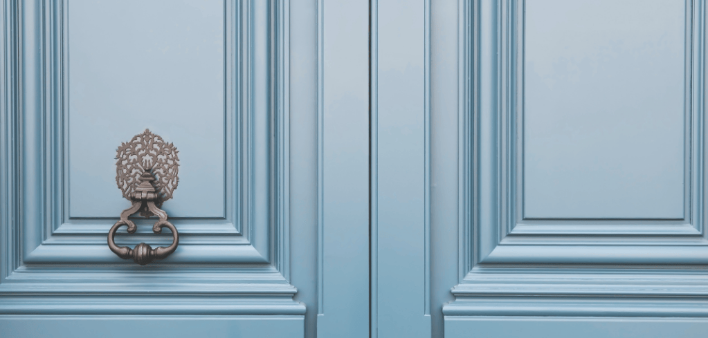 Ornate bronze door knocker with decorative backplate mounted on a light blue wooden door with paneled details.