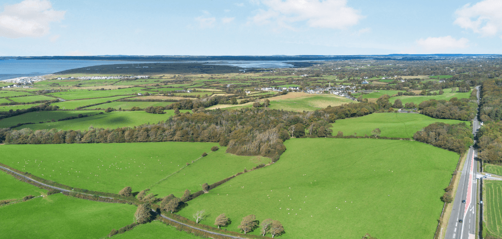 Aerial view of lush green farmland with hedgerows, a coastal bay, scattered trees, a village, and a highway on the right.