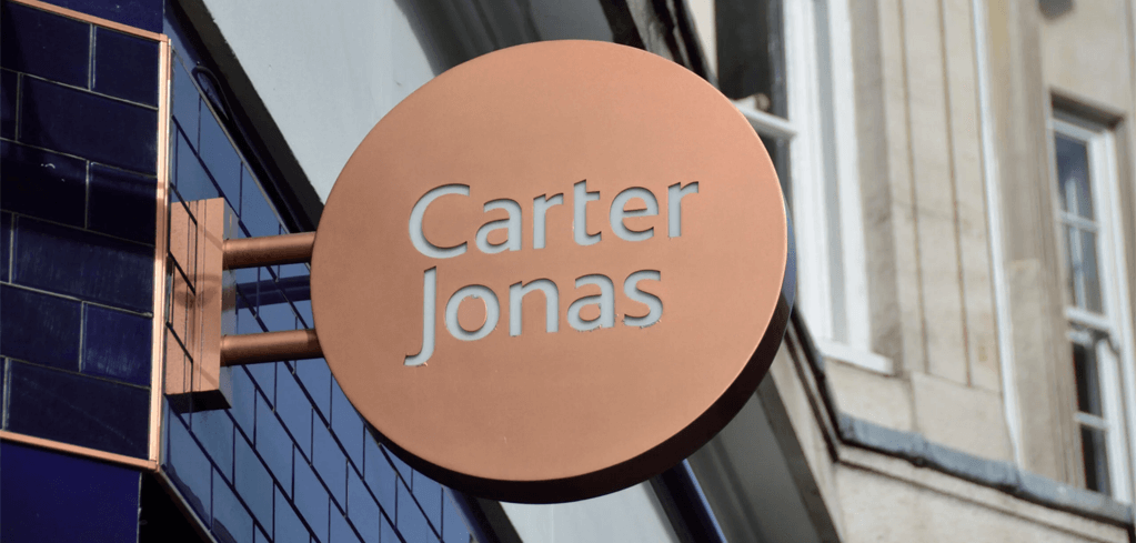 Copper circular sign with "Carter Jonas" text mounted on a building exterior with blue tiles and white facade.