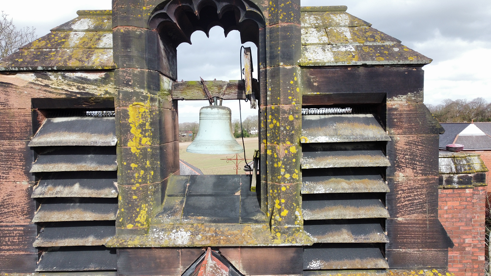 Large bell housed in an old stone bell tower with arched opening and weathered brick structure covered in moss.