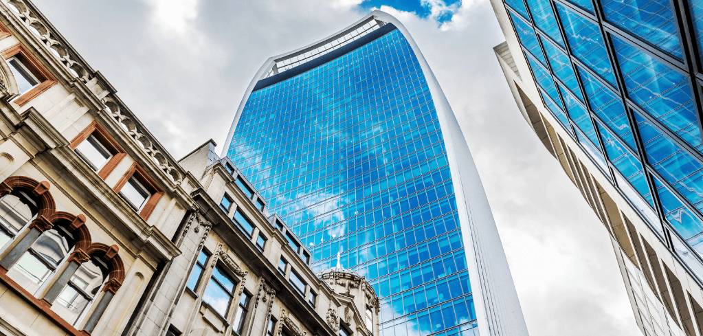 Looking up at London's Walkie-Talkie skyscraper, a curved blue glass tower, flanked by a classic stone building under a cloudy sky.