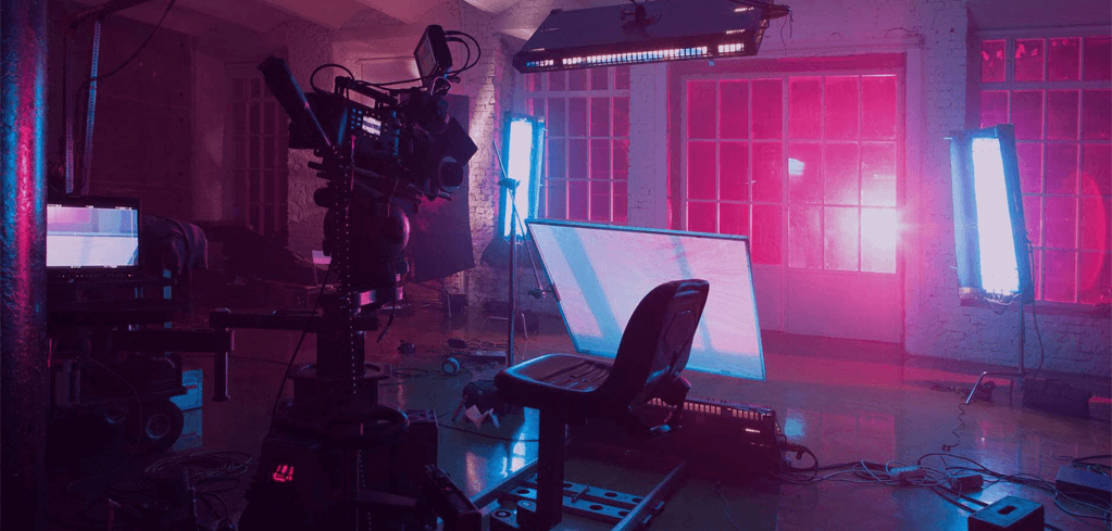 Film studio with professional camera, lighting equipment, and monitor bathed in vibrant pink and blue lighting.