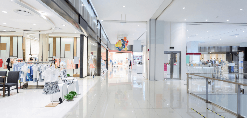 Modern shopping mall interior with bright white floors, clothing displays, and retail storefronts under recessed lighting.