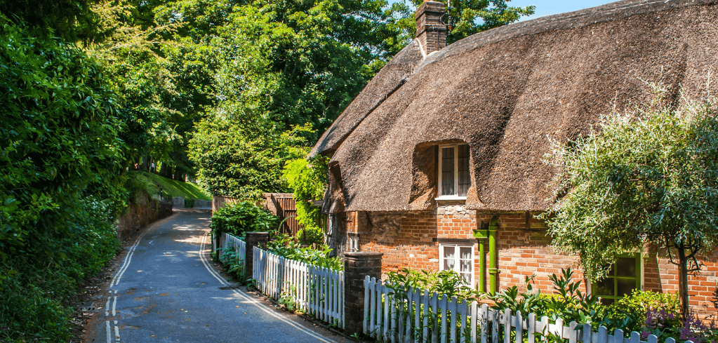 Traditional thatched cottage with brick walls and white picket fence along a narrow country lane surrounded by lush greenery.