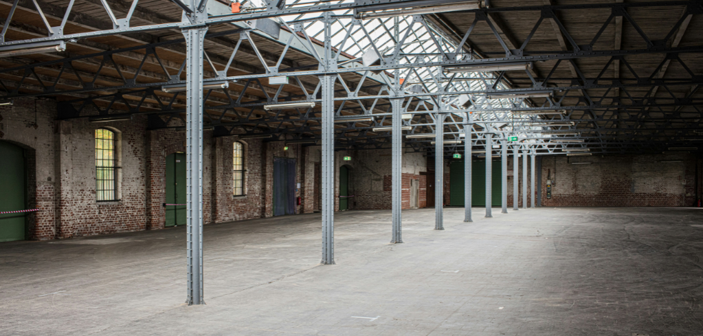 Large industrial warehouse interior with exposed brick walls, metal support columns, and a skylight ceiling in an empty space.