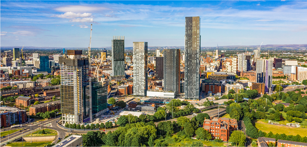 Aerial view of Manchester city skyline with modern skyscrapers, green parks, and surrounding urban landscape under blue sky.
