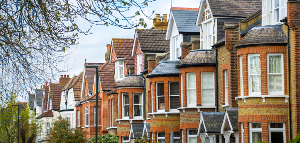 Row of traditional British terraced houses with red brick facades, bay windows, and distinctive rooflines against a cloudy sky.