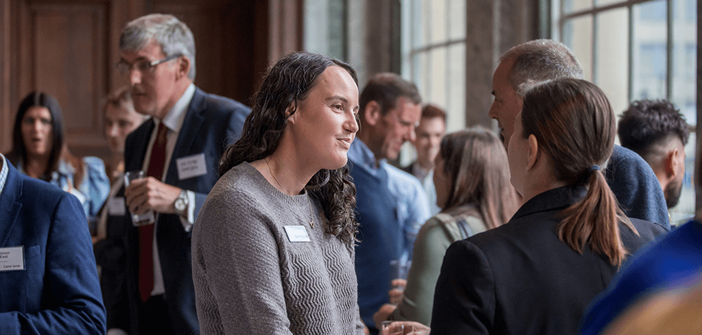 Professionals networking at a formal event in an elegant room with large windows, wearing name badges and business attire.