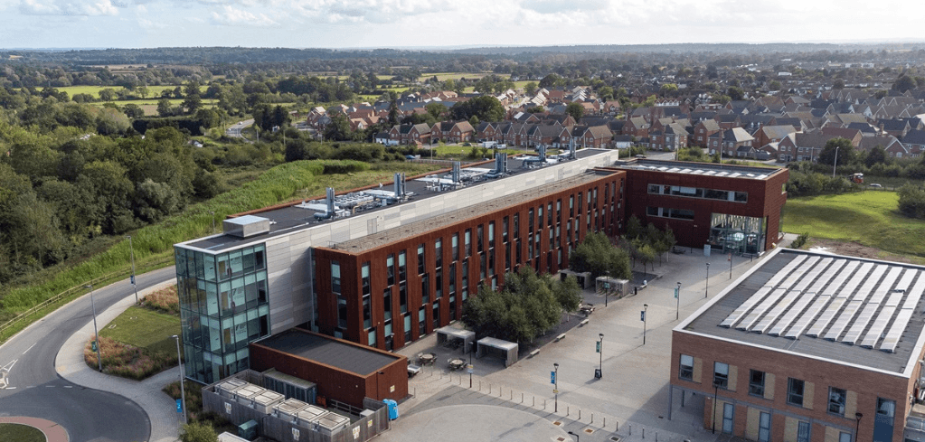 Aerial view of a modern red brick campus building with glass sections, surrounded by a plaza, greenery, and a residential neighborhood.