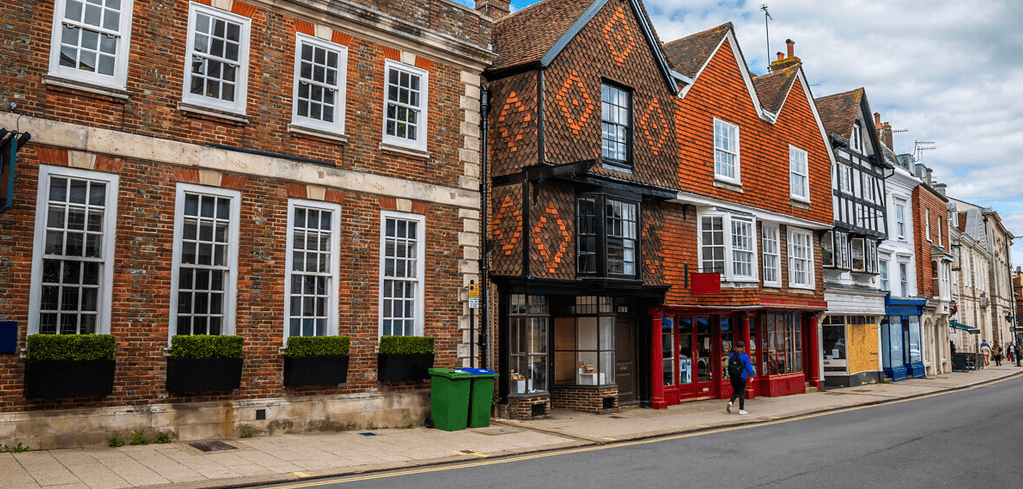 Historic English street with brick buildings, Tudor-style architecture, and colorful storefronts under a cloudy sky.