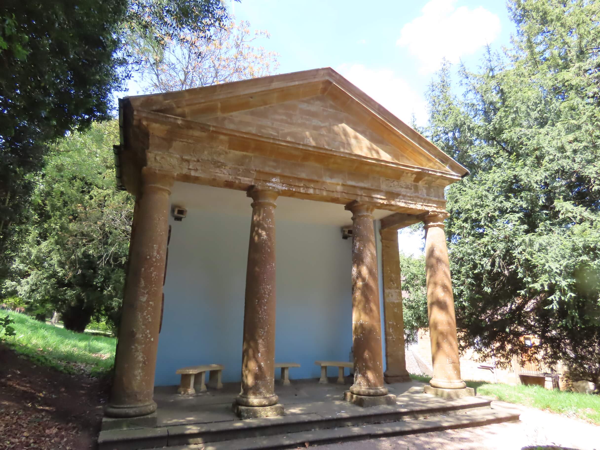 A small classical stone pavilion with four columns and a triangular pediment, surrounded by lush green trees in a park.