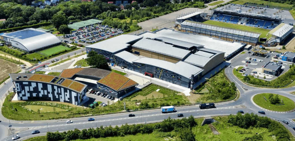 Aerial view of a large sports and leisure complex with a stadium, arena, green rooftops, parking lots, and surrounding roads.