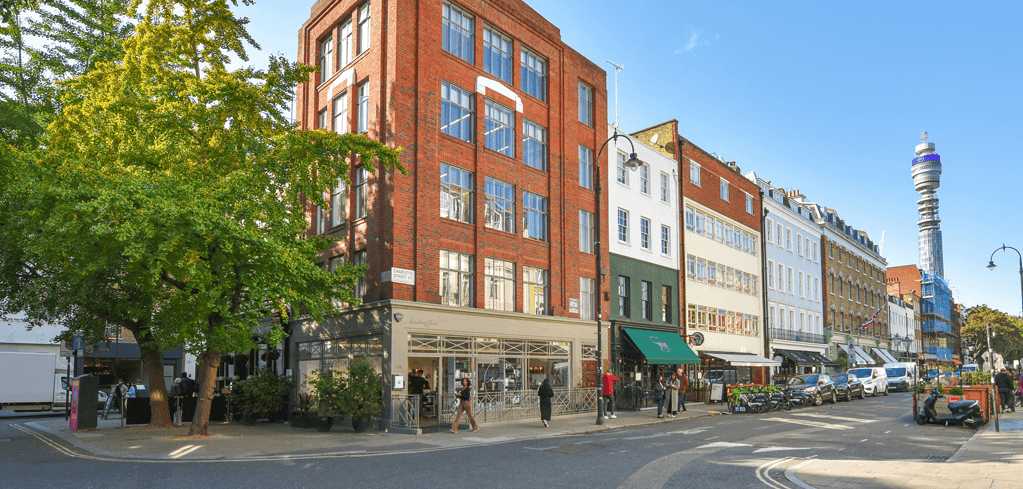 A sunny London street with red brick buildings, shops, pedestrians, a green tree, and the BT Tower visible in the background.