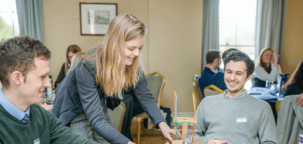 People in a workshop setting collaborating on a small colorful structure made of sticks in a bright meeting room.