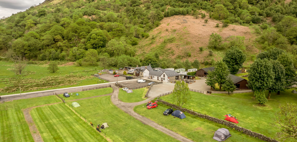 Aerial view of a rural campsite with tents and cars on green fields, nestled at the base of a lush green hillside.